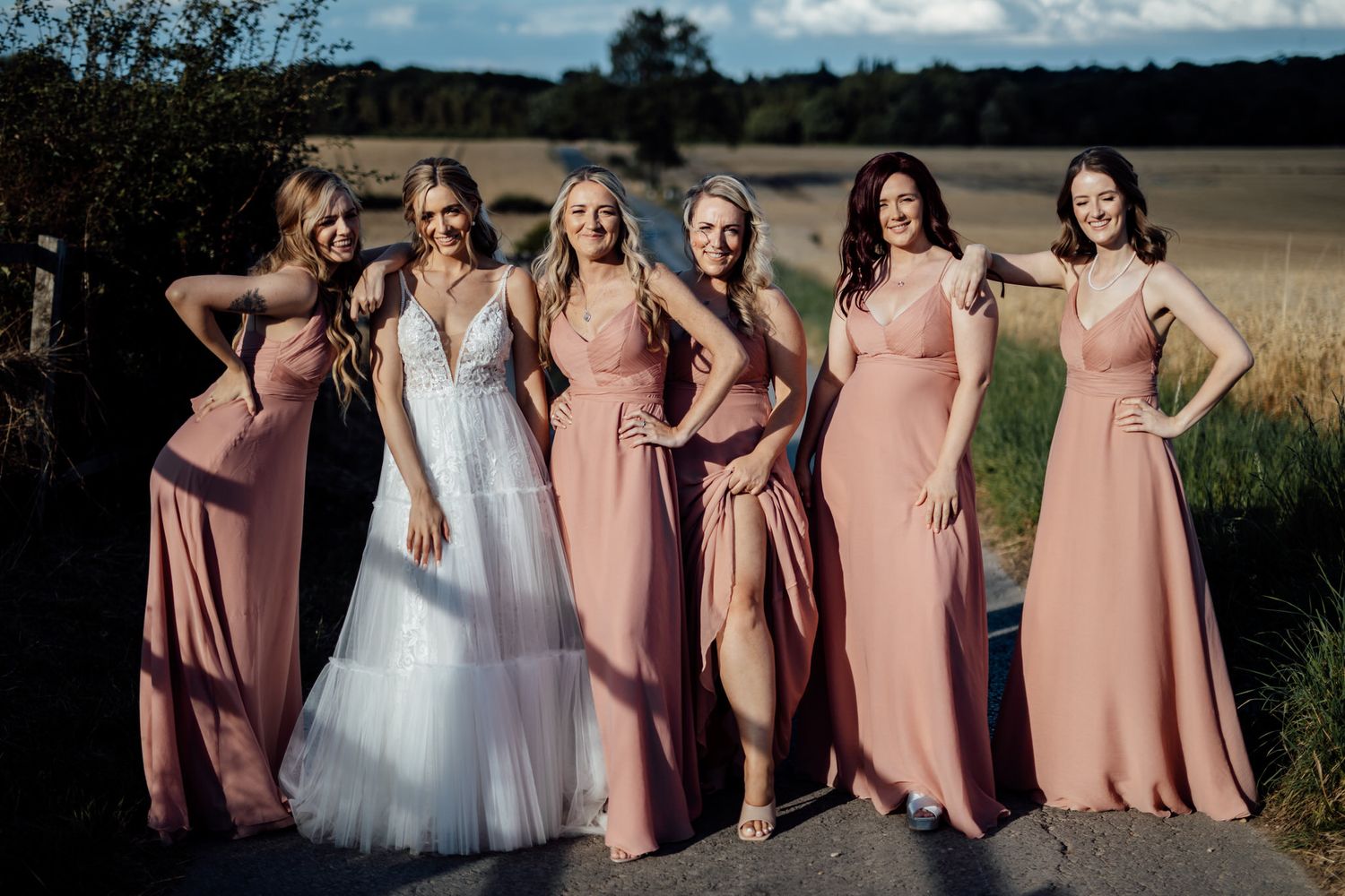 Bridesmaids in pink dresses pose playfully with bride in white gown against golden field backdrop.
