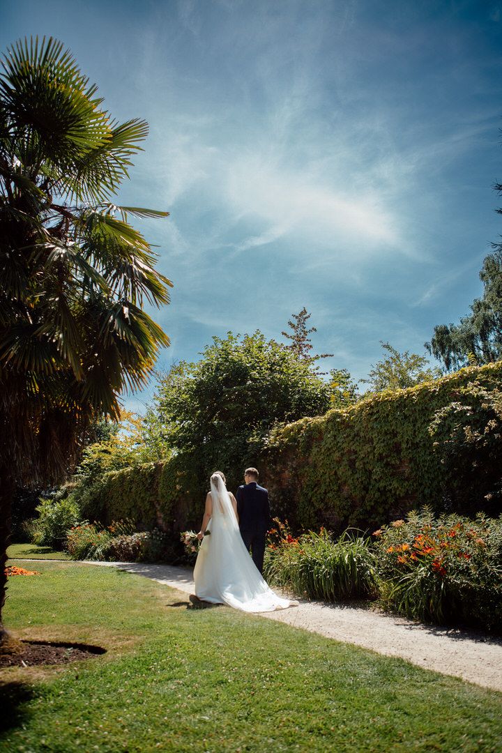 Wedding couple walks along garden path under blue sky with palm trees and lush greenery.