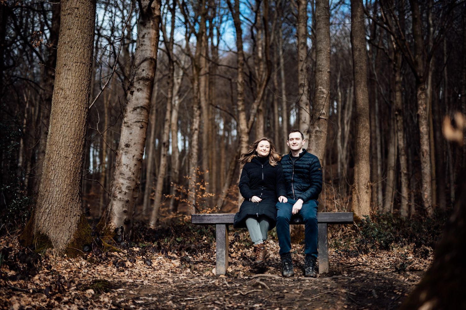 Two people sitting together on a bench surrounded by bare trees in a forest setting.