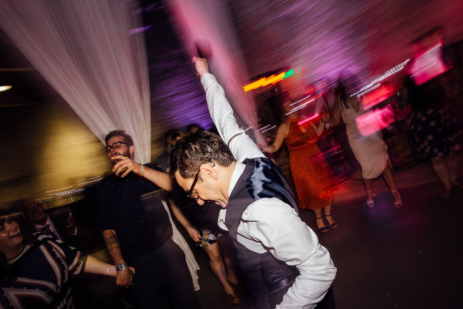 Wedding party dancing energetically under red and purple lighting effects.