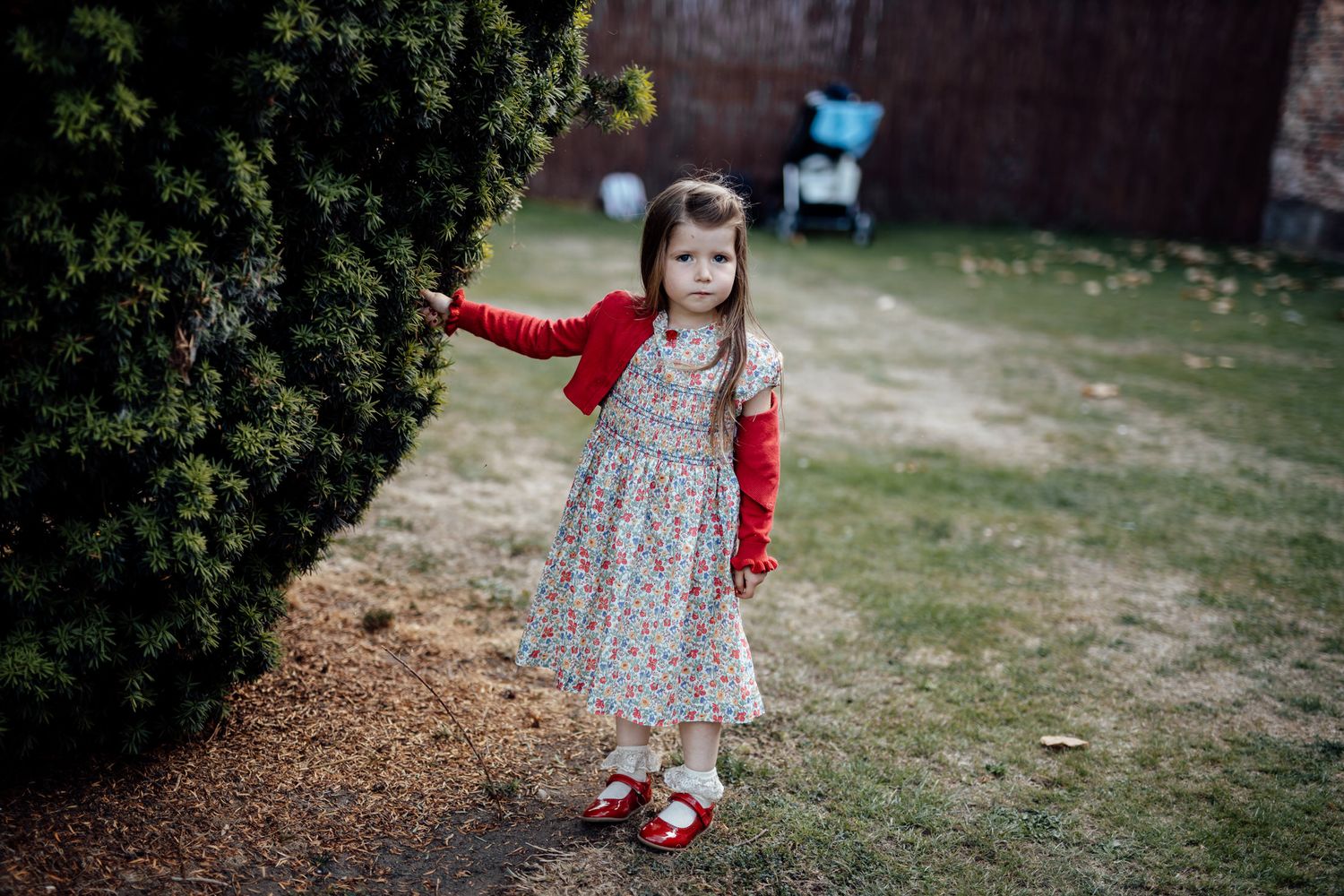 Little girl in a floral dress and red shoes walking through a garden path.