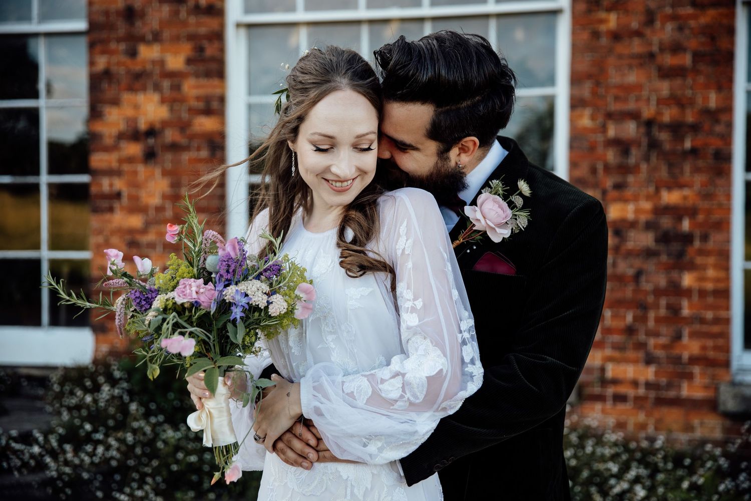 A couple in wedding attire share intimate moments in front of a brick building with a bouquet of purple flowers.