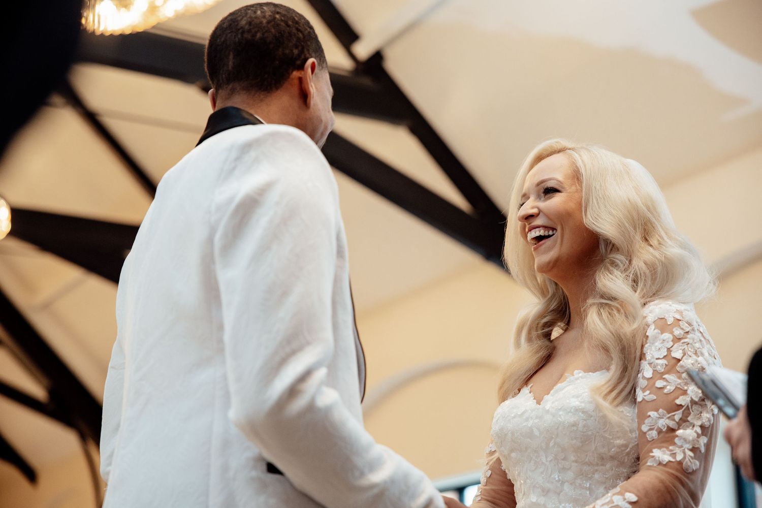 Radiant blonde bride smiling joyfully at her groom during an elegant indoor wedding ceremony.