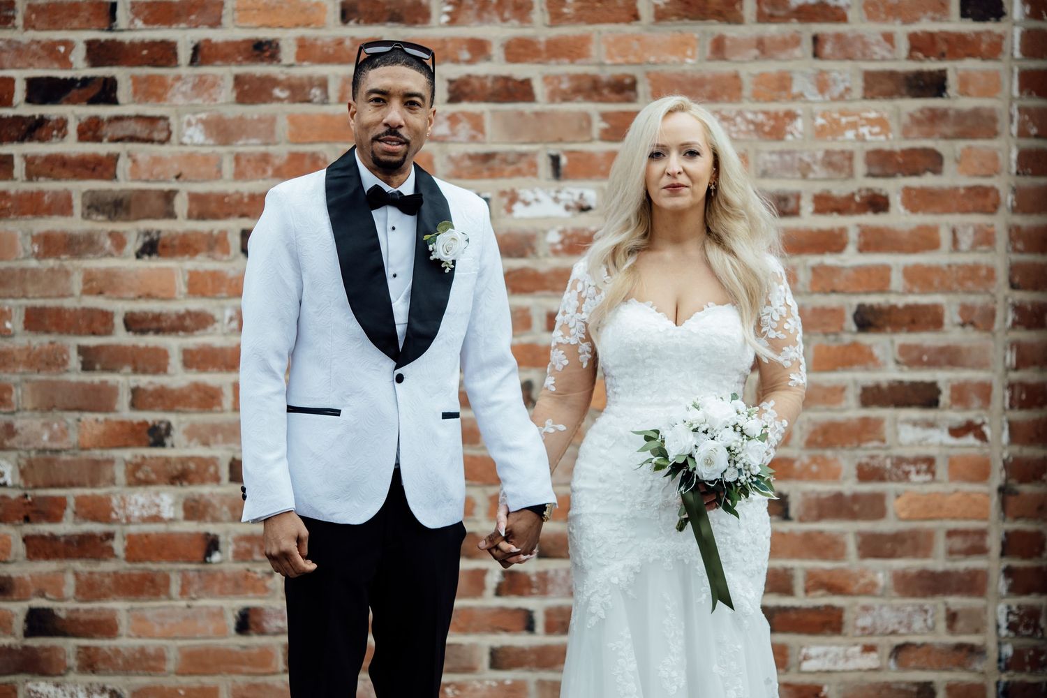 Bride and groom holding hands in front of brick wall, groom in white tuxedo, bride in lace gown.