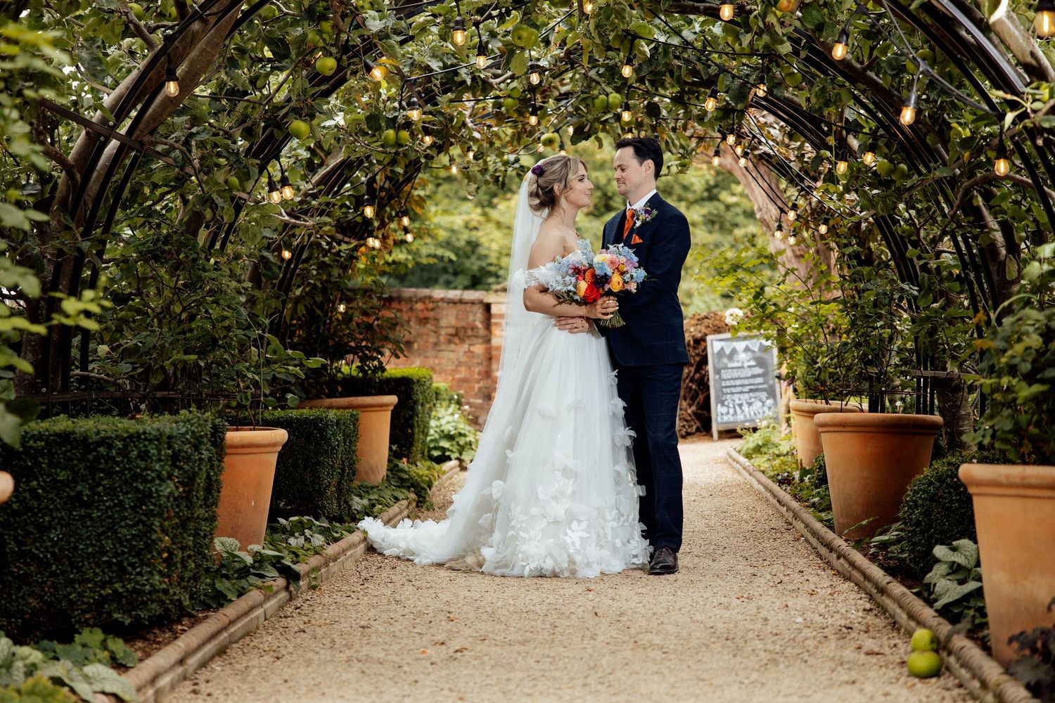 Wedding portraits taken in a garden pathway with brick walls and string lights creating a romantic atmosphere.