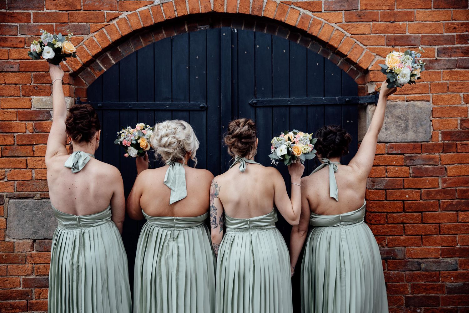 Bridesmaids in sage green dresses standing in a row against a brick wall holding white and peach bouquets.