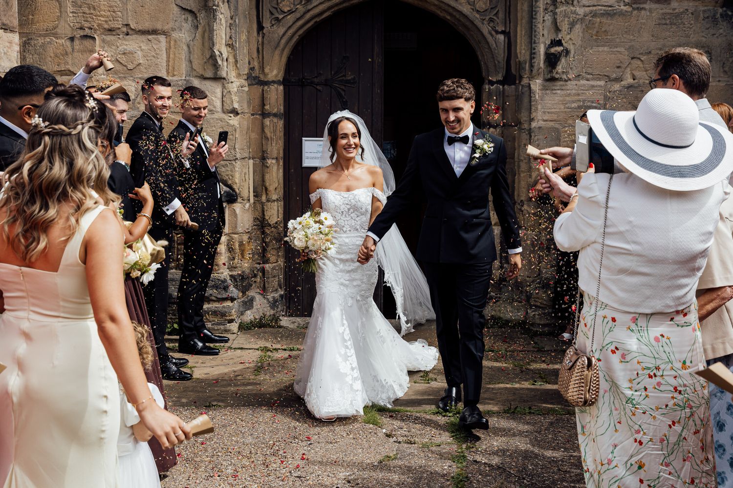Wedding guests celebrate with confetti outside a historic stone church building.