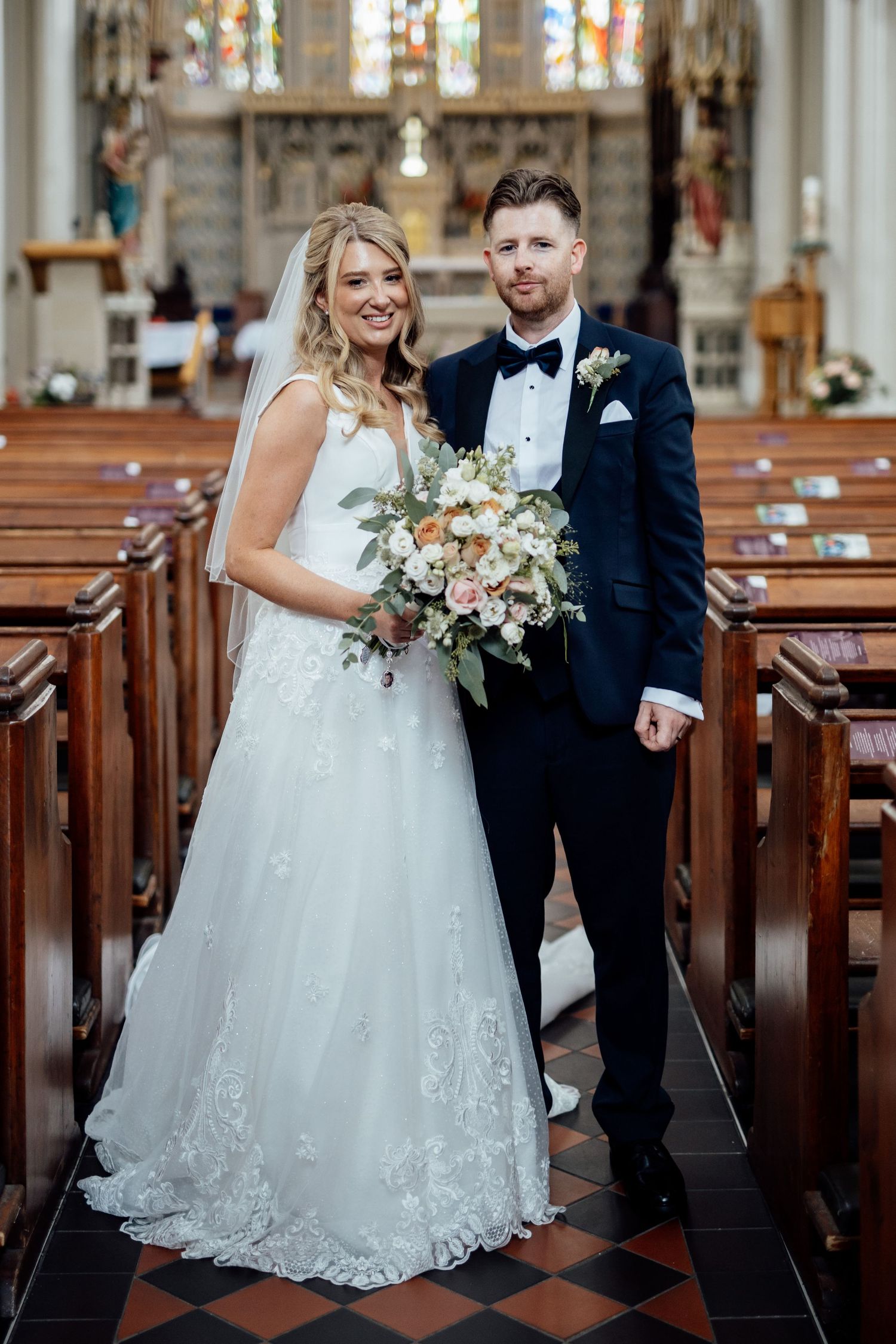 Wedding couple standing in a grand church with stained glass windows and wooden pews.