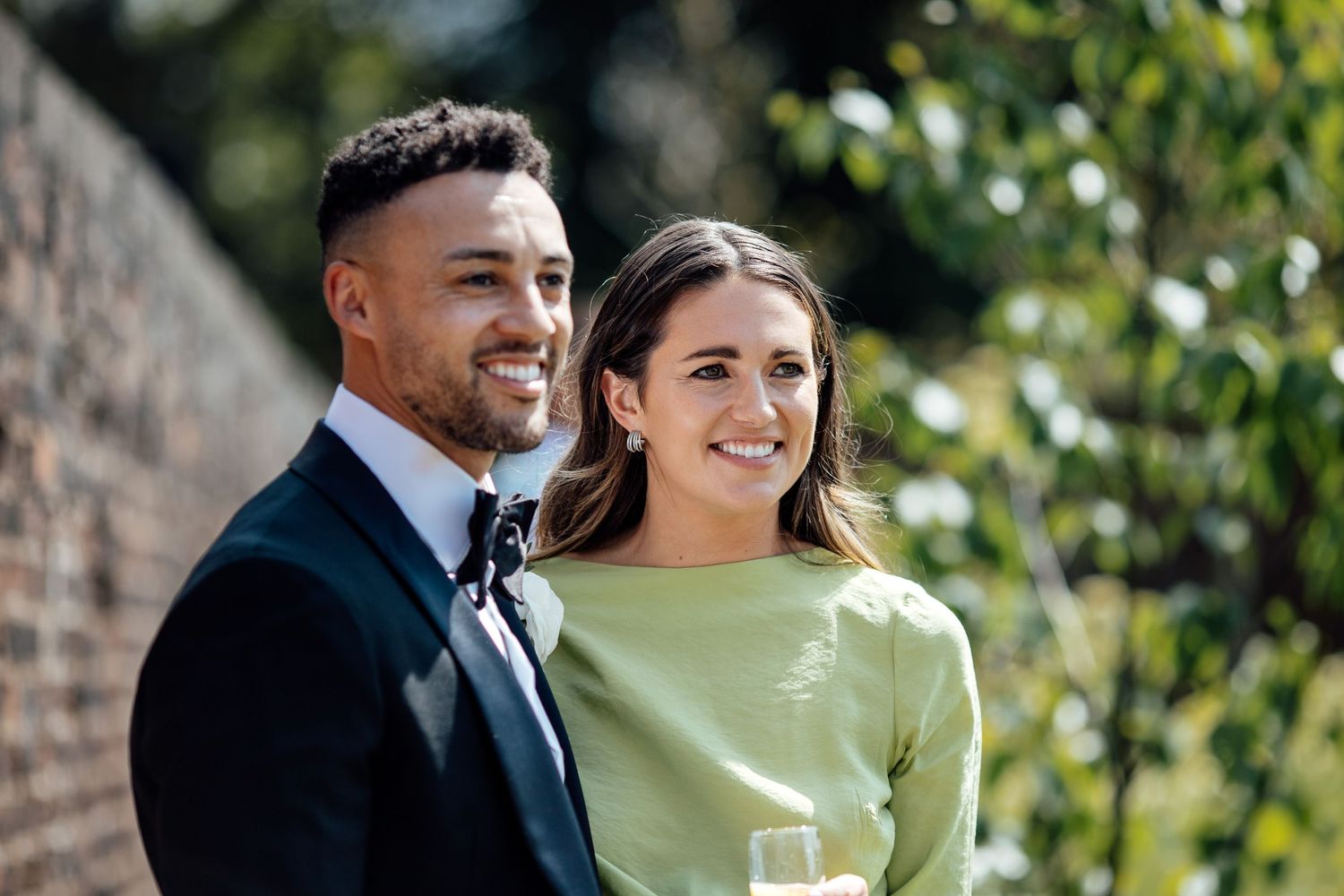 Couple in formal attire laughs together against a sunlit garden backdrop with greenery.