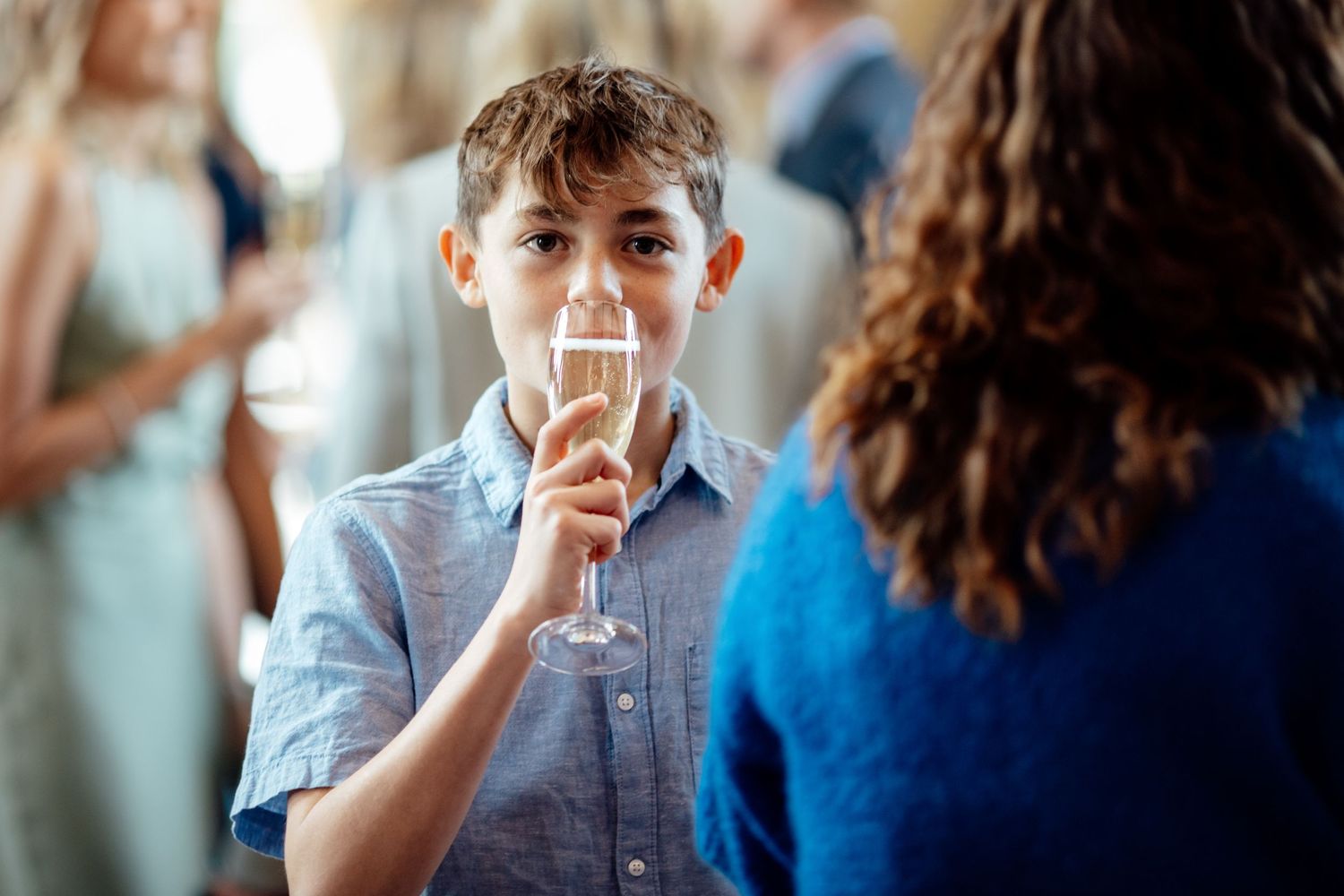 Young guest in blue shirt sipping champagne during wedding reception.