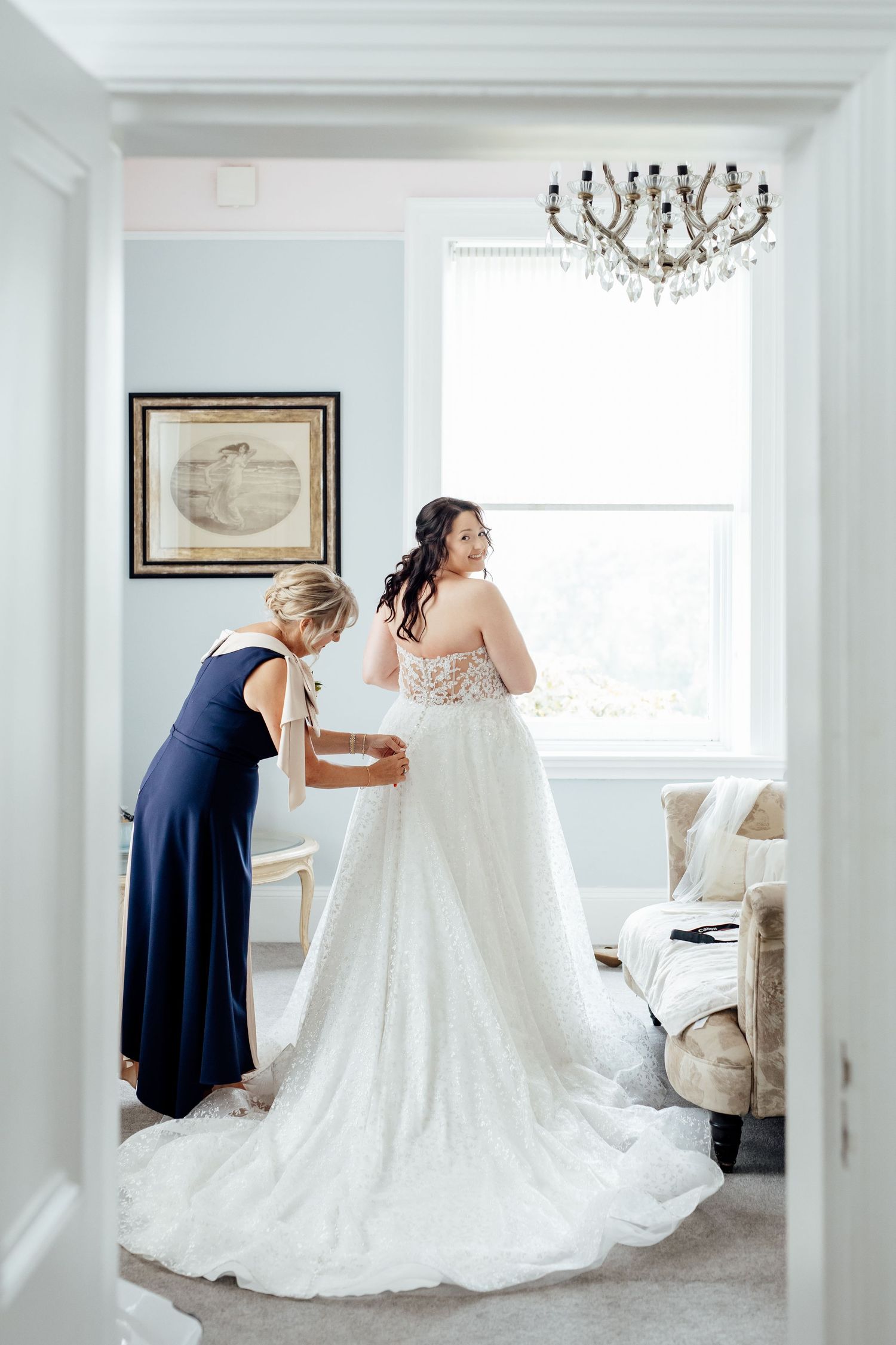 Wedding dress preparation in bright white room with chandelier and elegant decor.