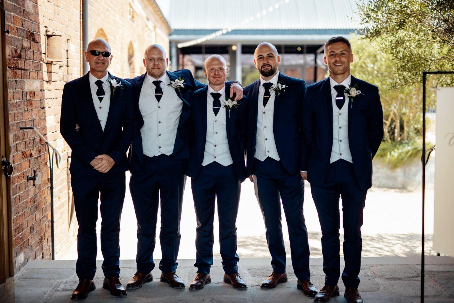 Groomsmen in formal black tuxedos pose together for wedding party photo.