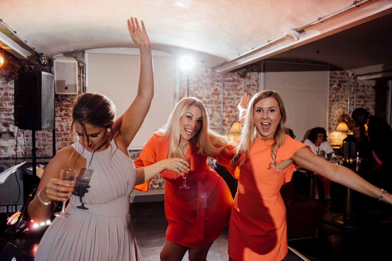 Three wedding guests in bright orange and white outfits dancing enthusiastically at a reception.