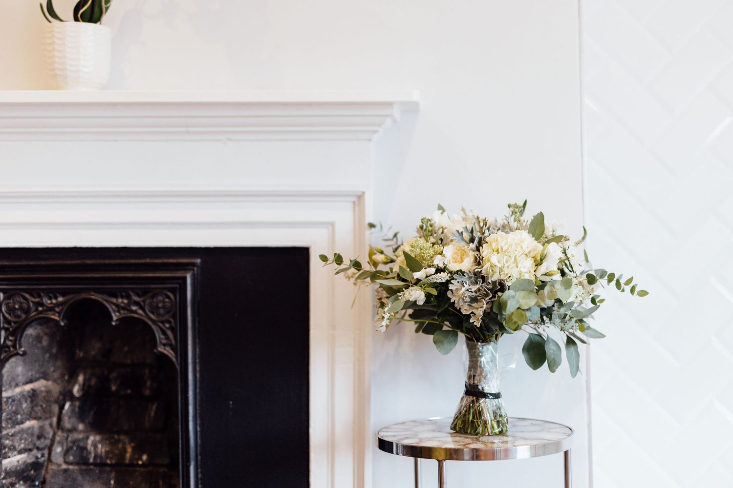 Elegant white floral arrangement with eucalyptus in a vase beside a fireplace.