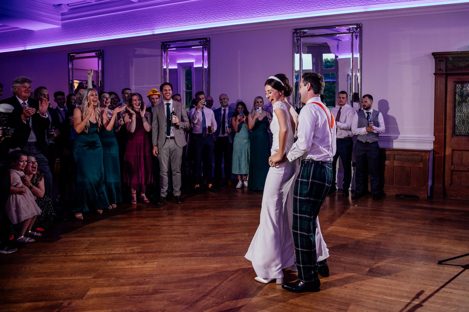 Wedding party dancing under purple lighting in a spacious ballroom.