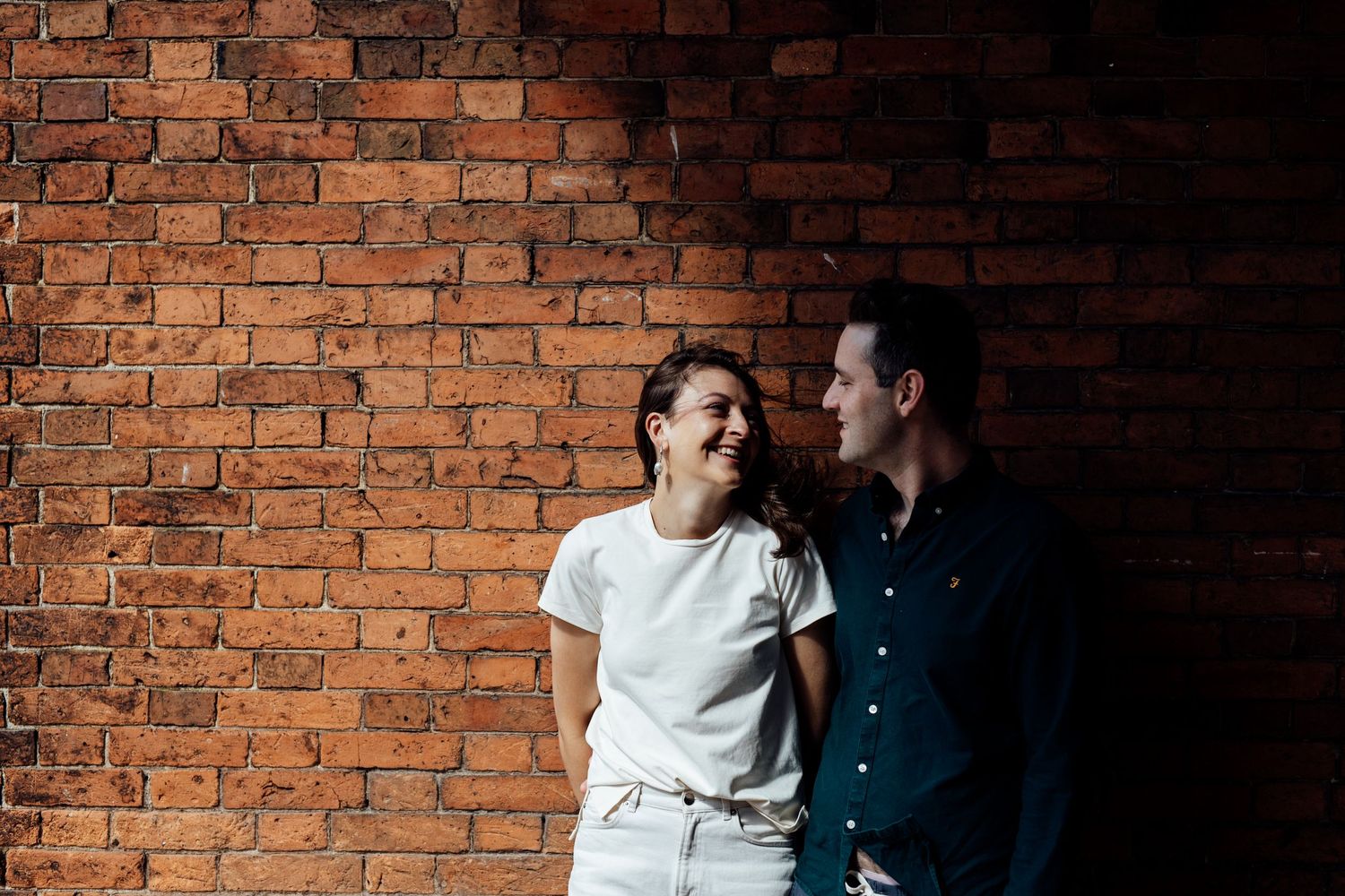 A couple shares intimate moments against a textured brick wall with dramatic lighting.