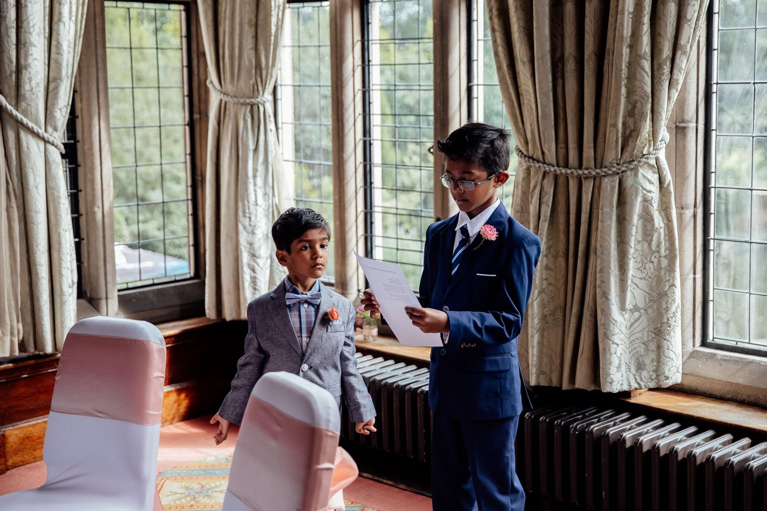 Two boys in matching navy suits share a special moment in an elegant room with draped windows.