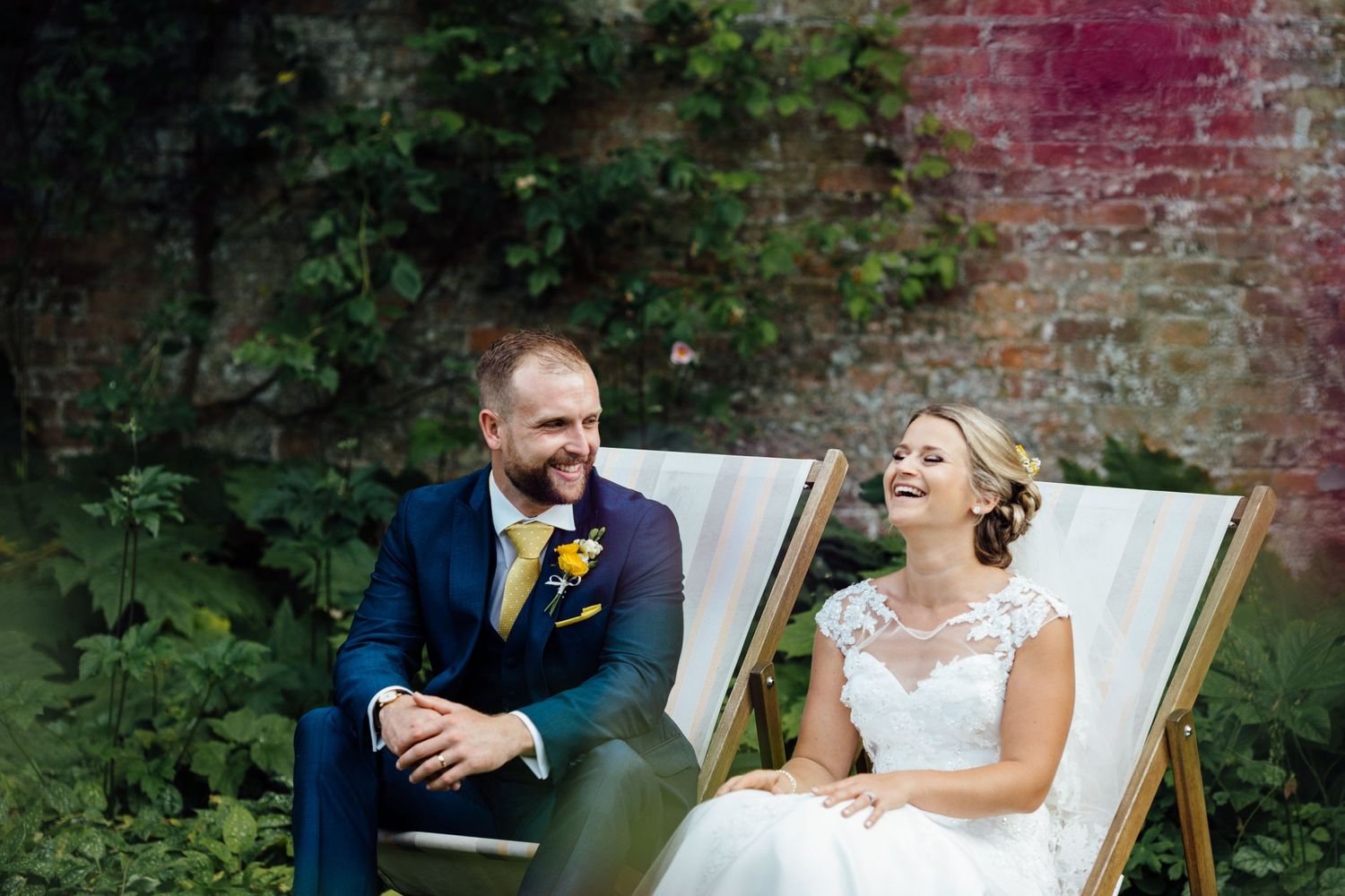 Couple relaxing in white deck chairs in a lush garden setting surrounded by brick walls.