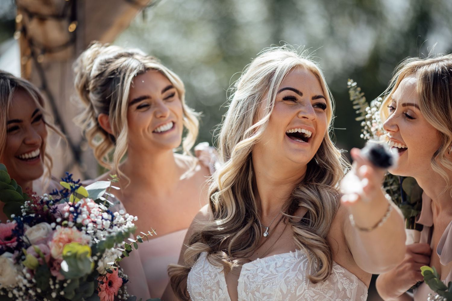 Joyful bride in lace gown laughing with bridesmaids holding colorful bouquets at a sunny outdoor wedding.