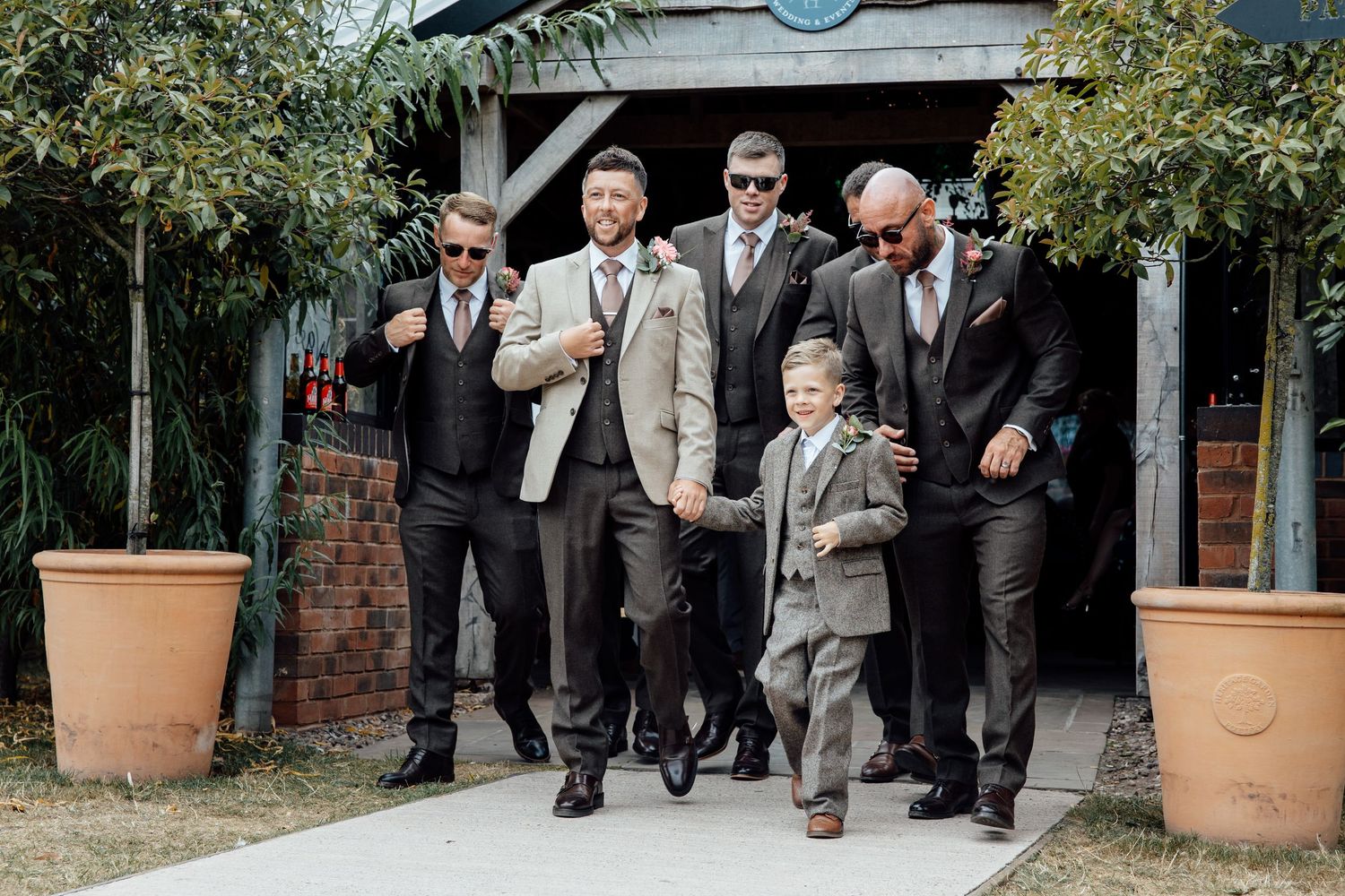 Groomsmen and ring bearer walking confidently in suits and sunglasses at an outdoor wedding venue.