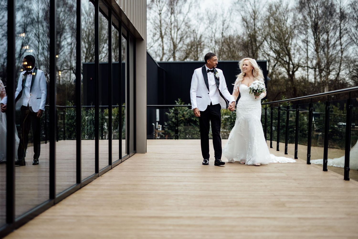 A bride and groom walk hand in hand along a modern outdoor deck at their wedding venue.