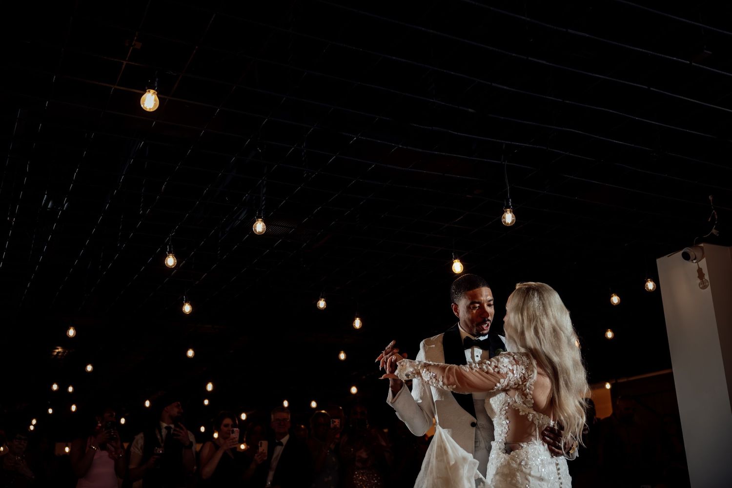 Bride and groom share first dance under warm Edison bulb lights at dark romantic wedding reception.