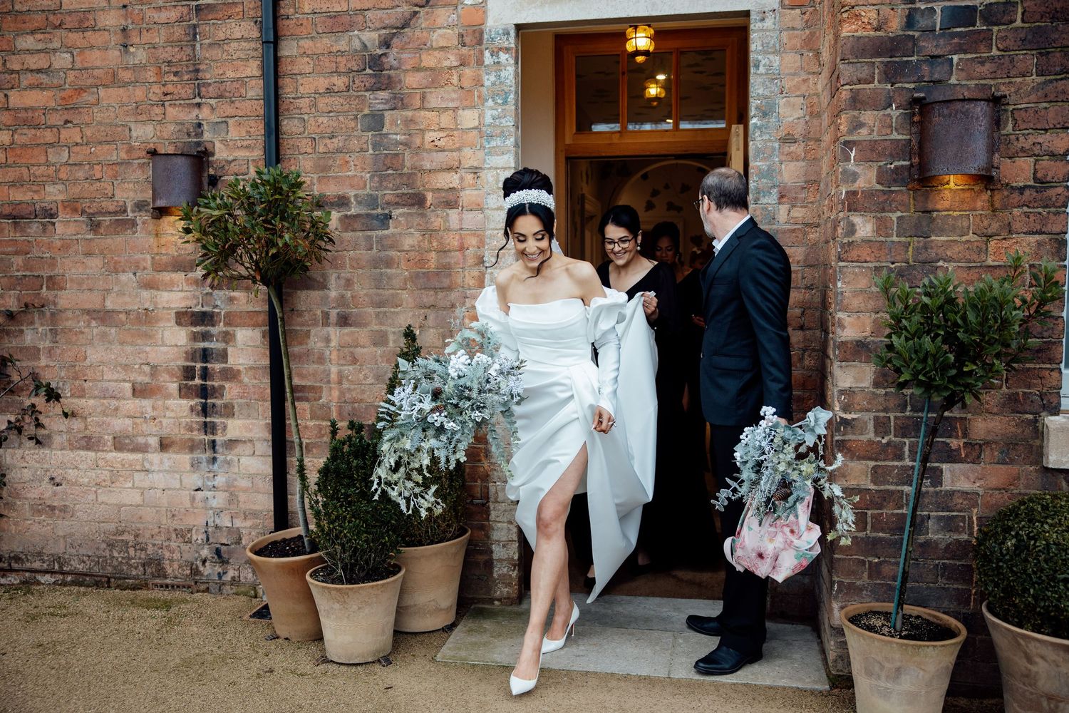 Wedding party enters brick venue entrance decorated with potted plants.