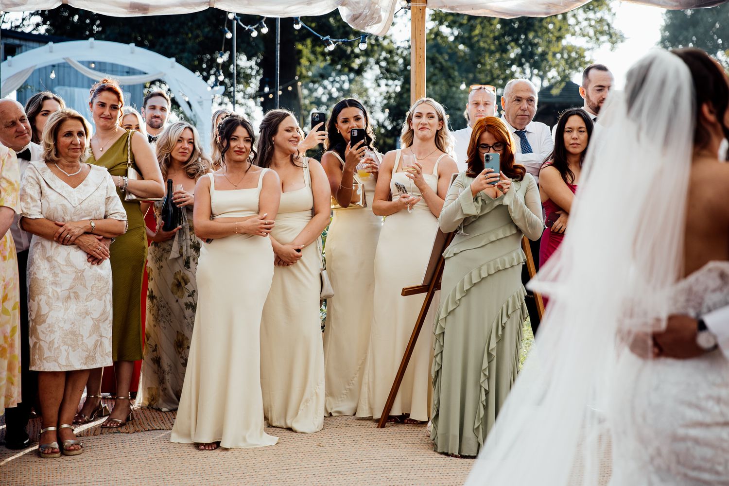 Wedding party in neutral dresses poses together outdoors under white tent.
