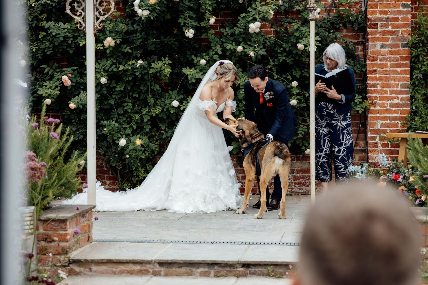Wedding moment with a friendly dog at a brick-walled garden entrance.