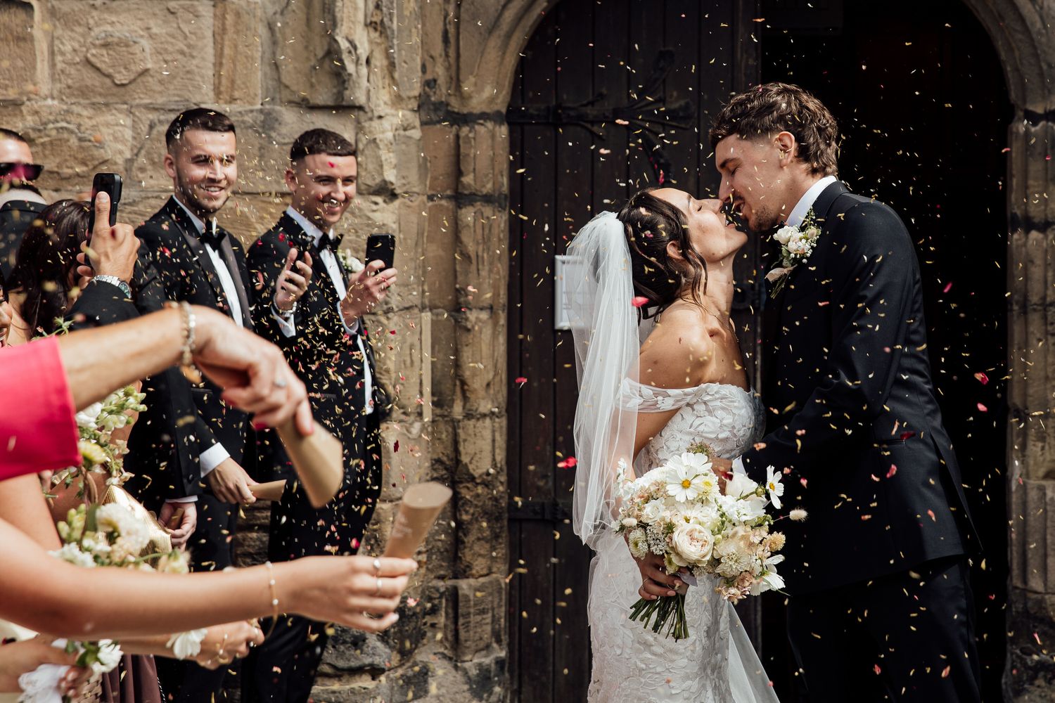 Wedding ceremony guests throw confetti outside historic stone church.