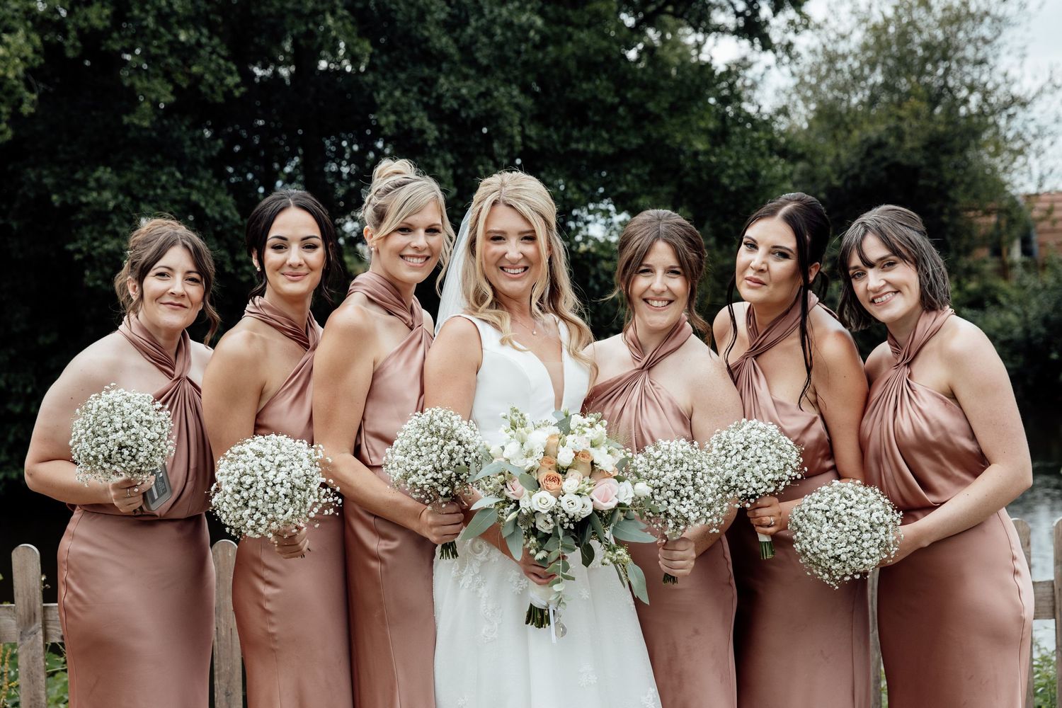 Bridal party in blush dresses holding white bouquets outdoors.