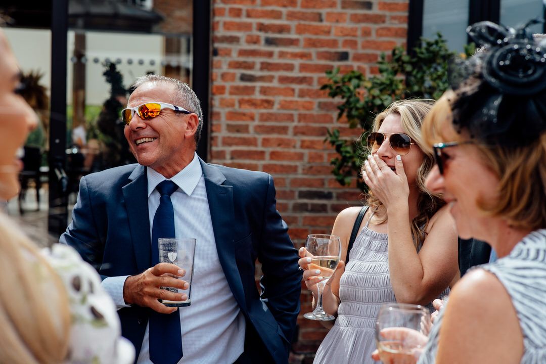 Wedding guests enjoying drinks and conversation at an outdoor reception with brick wall backdrop.