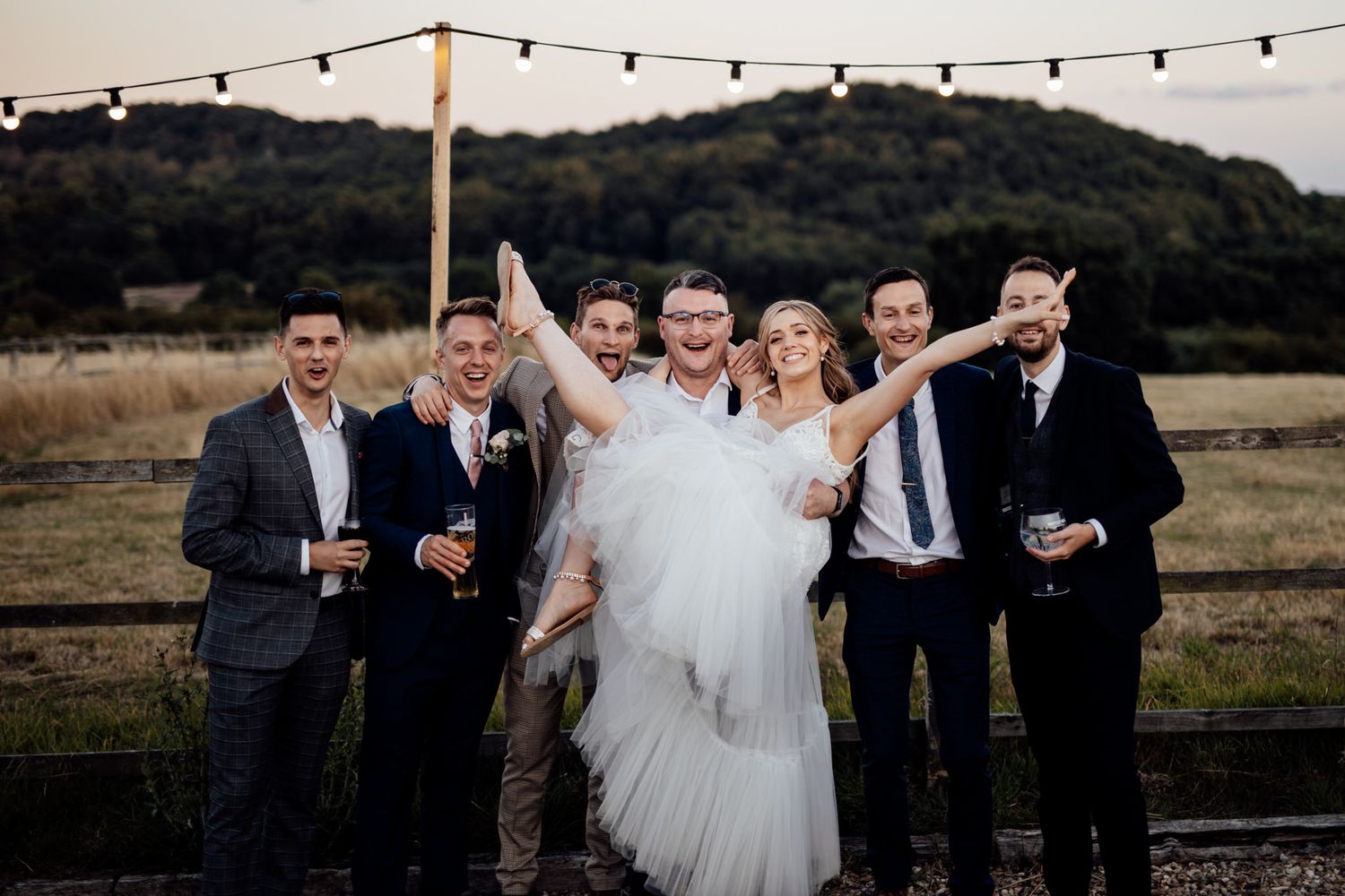 Wedding celebration group poses under string lights at dusk with mountains in background.