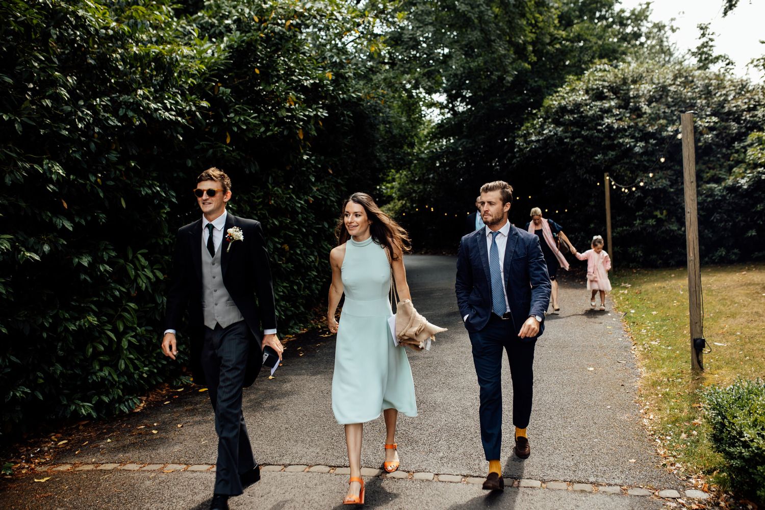 Wedding guests walking along tree-lined pathway at outdoor venue.