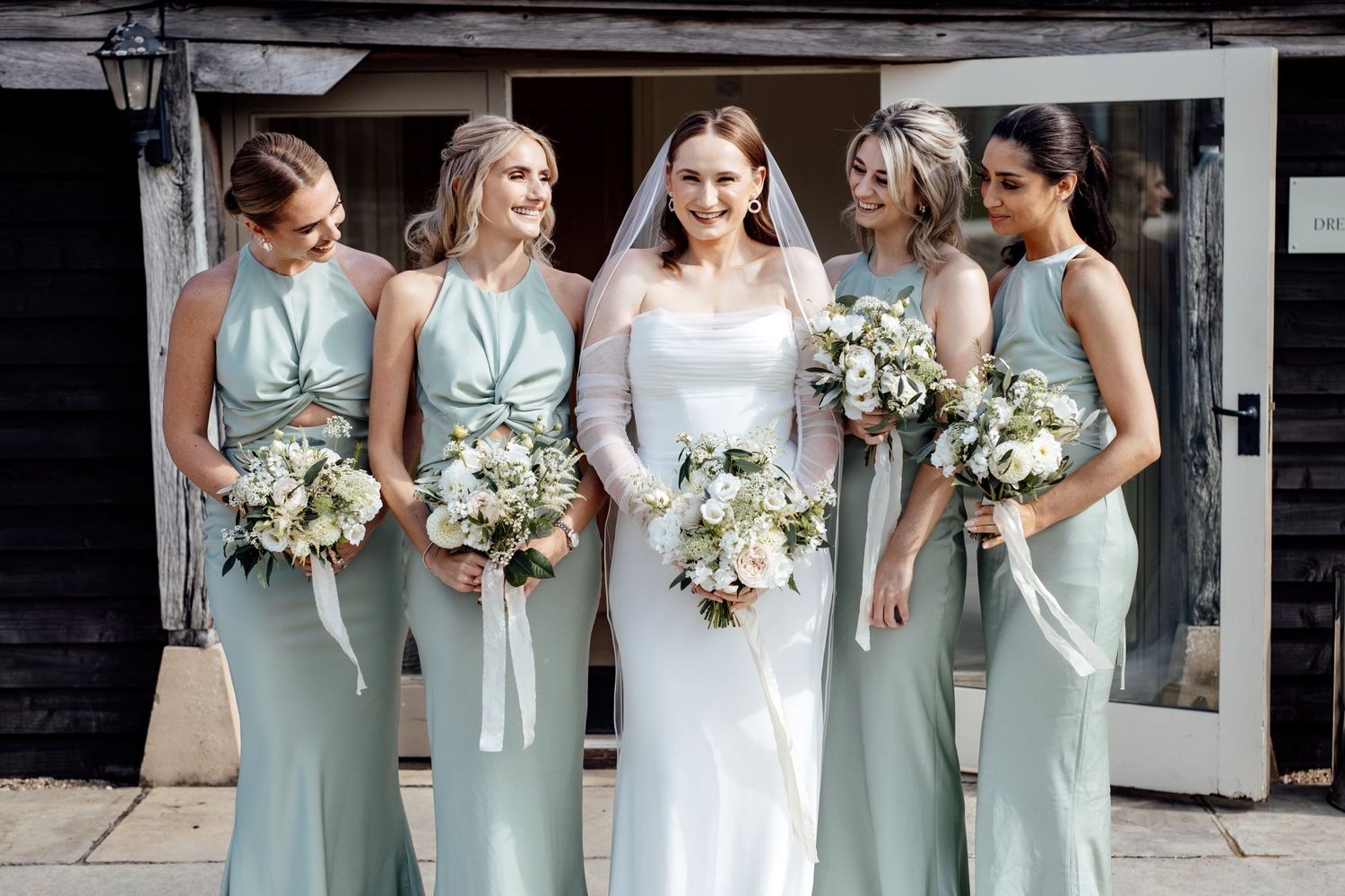 Wedding party poses together wearing sage green and white attire against rustic backdrop.