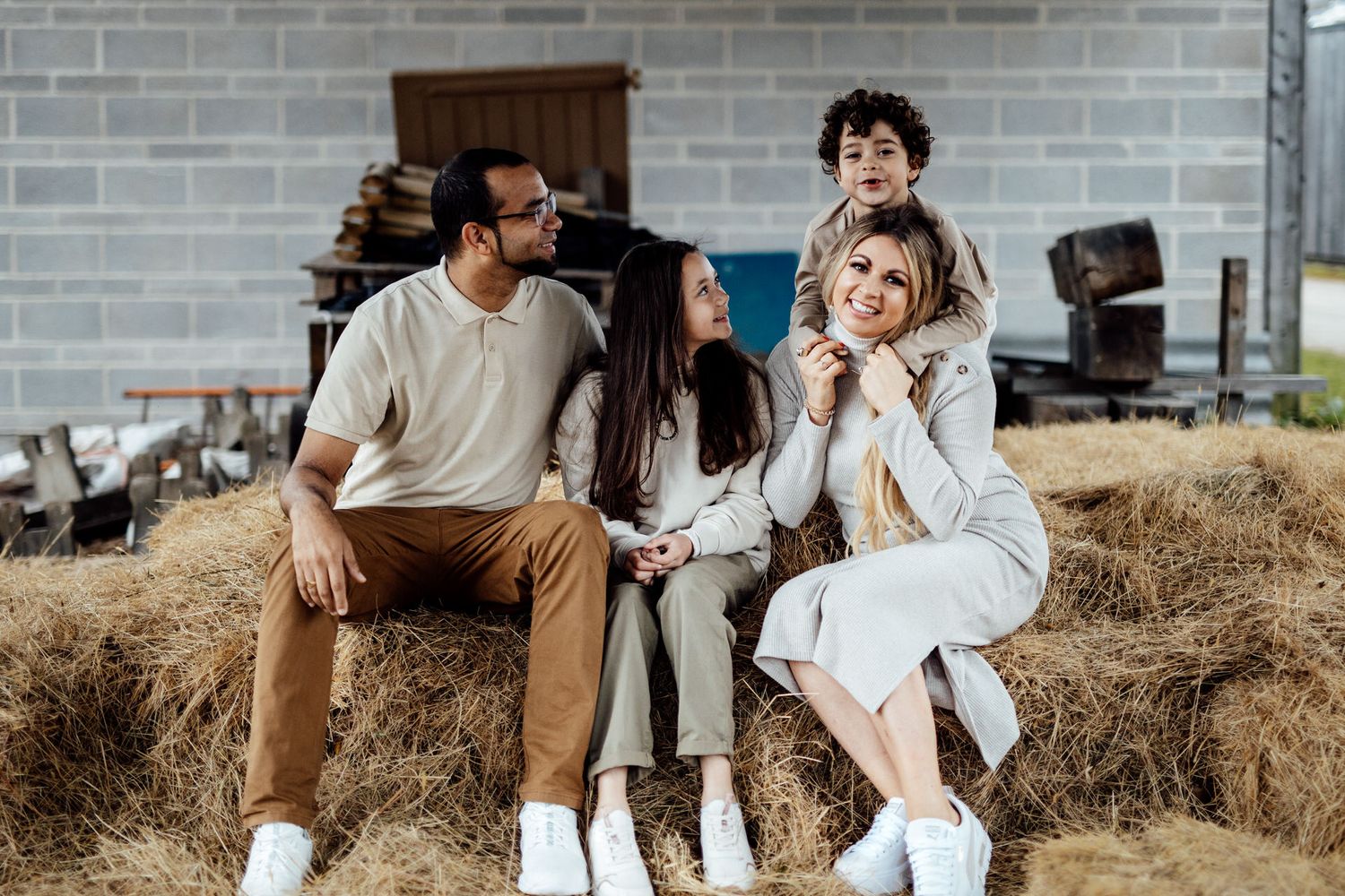 Candid family moment captured on rustic hay bales against a brick wall backdrop.