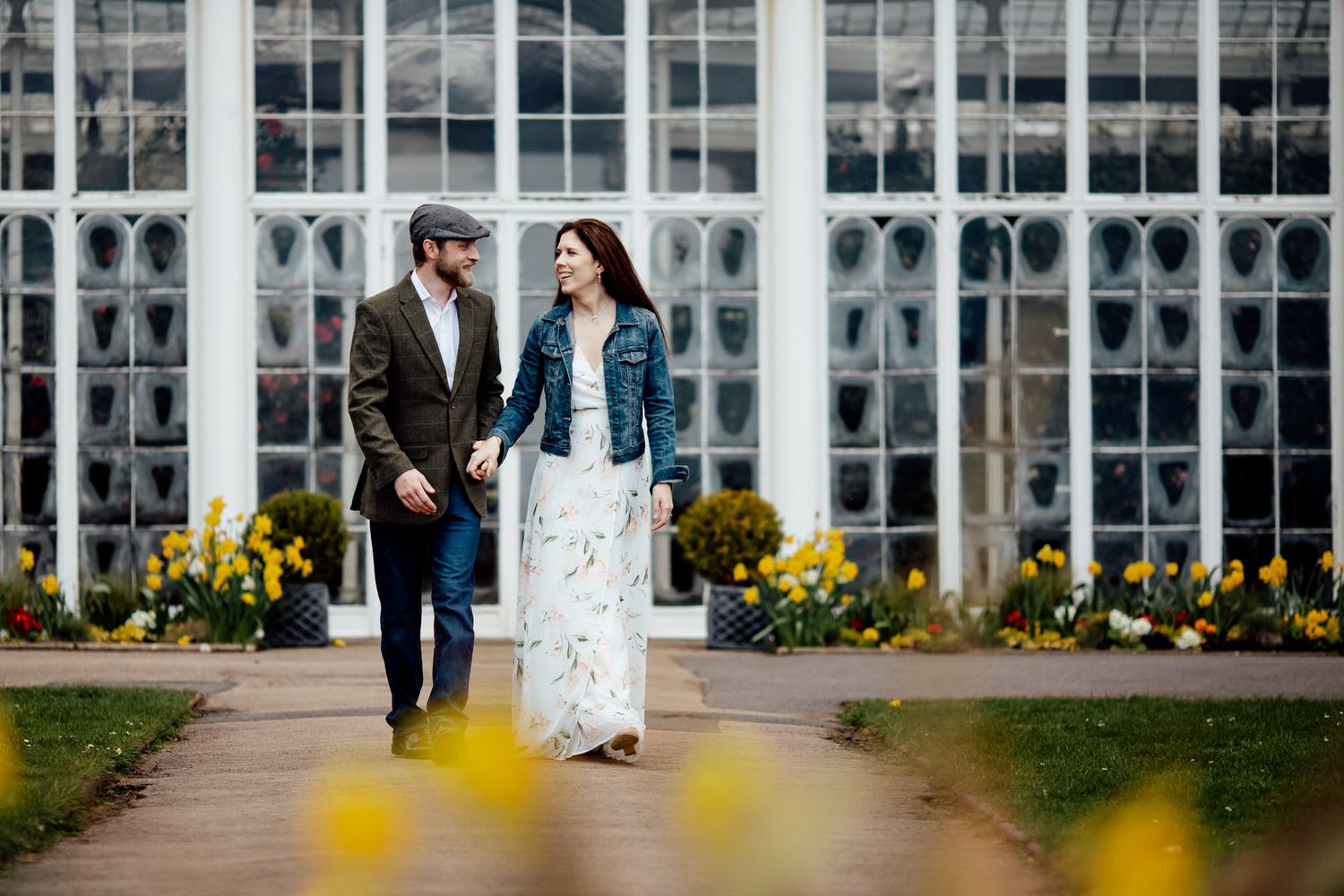 A couple stands together on a path in front of a grand conservatory with yellow flowers.