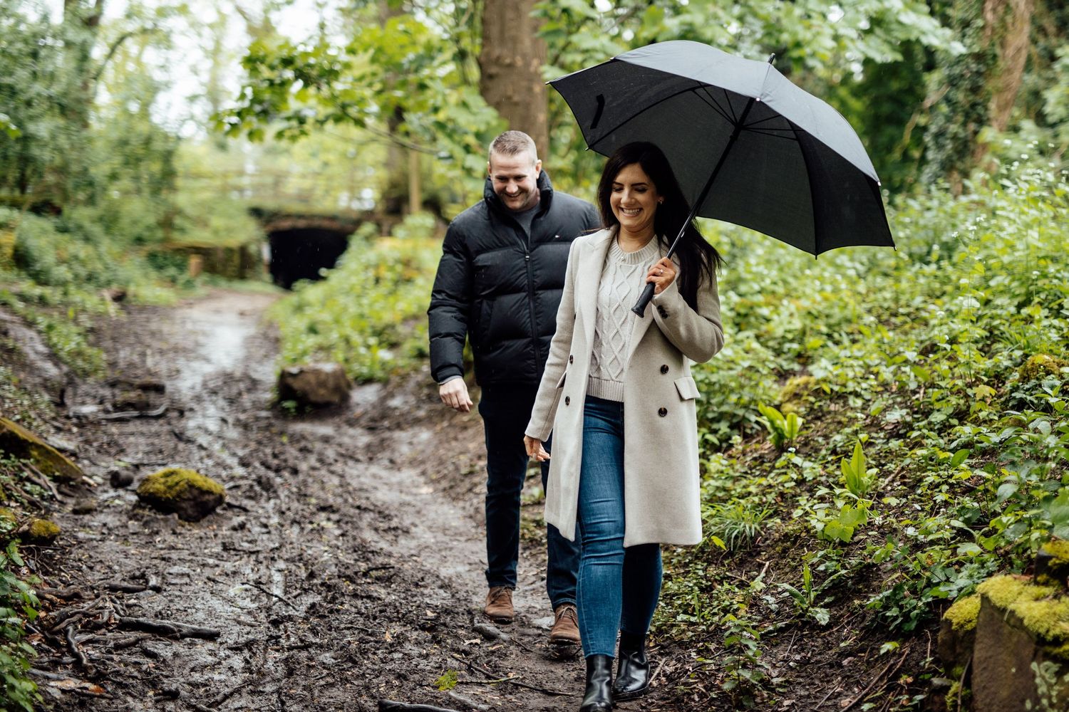 A couple walks along a muddy forest trail with a black umbrella on a rainy day.