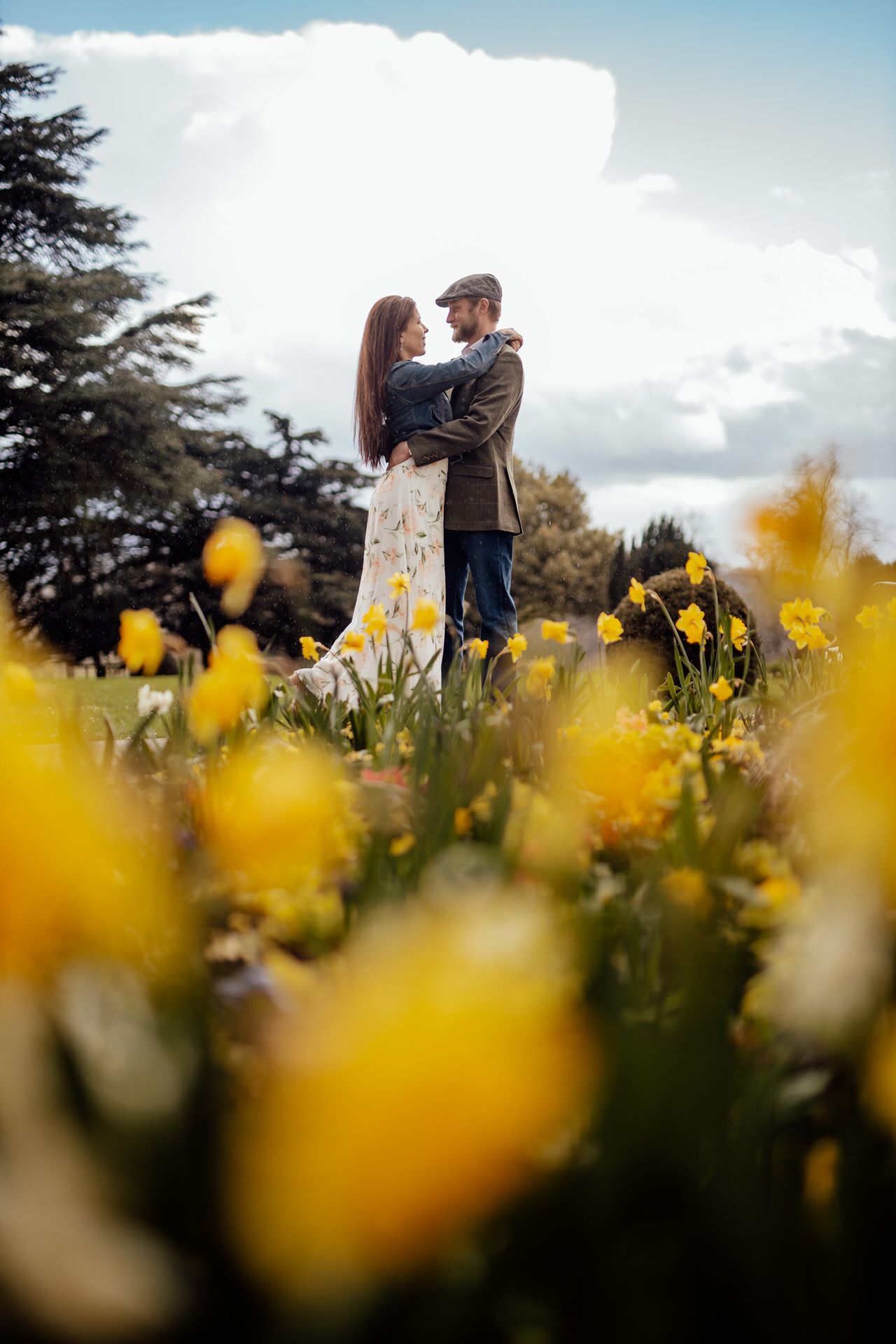 Couple embracing in field of yellow daffodils under bright blue sky.