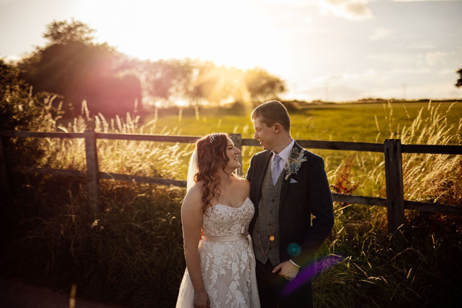 A couple stands together in golden sunset light near a rustic fence.
