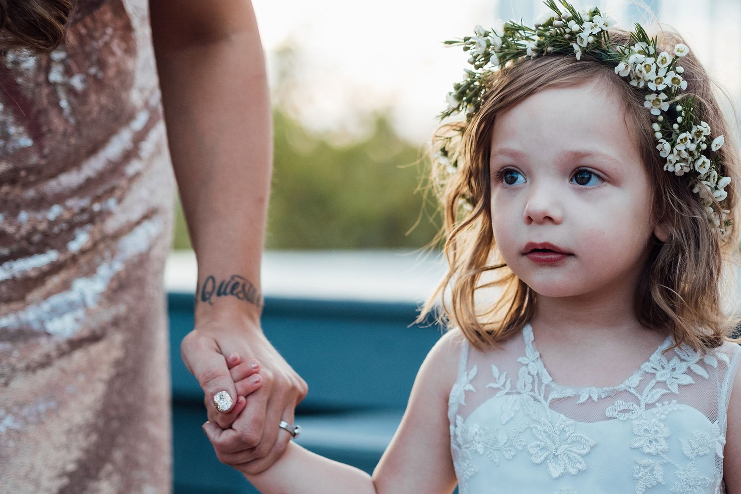 A young flower girl wearing a delicate white floral crown and dress smiles during an outdoor wedding ceremony.