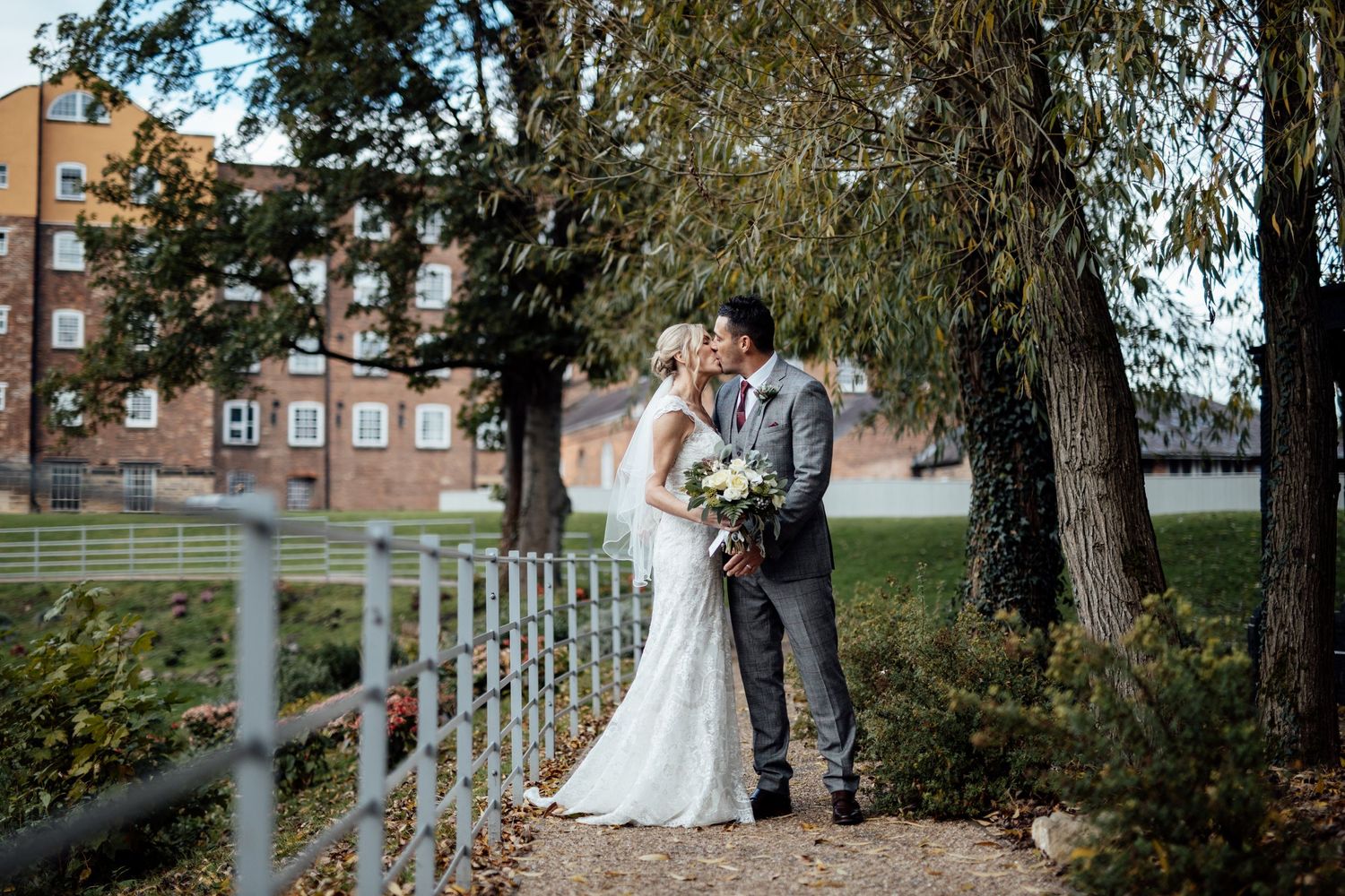 Newlyweds sharing intimate moments along a countryside fence with trees in background.