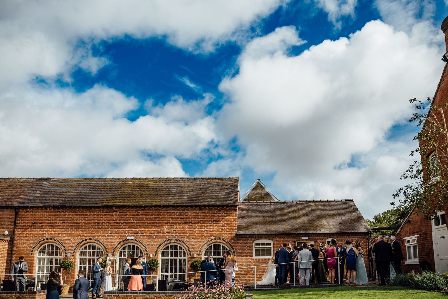 Panoramic view of a historic brick venue with wedding guests on the grounds.