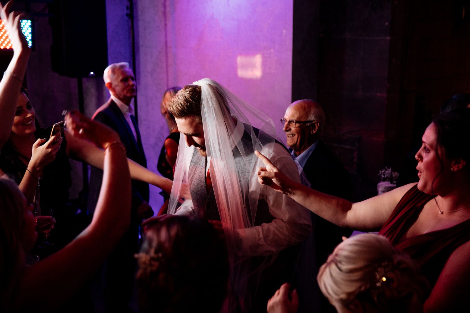 Wedding guests dance and celebrate on a dimly lit dance floor with purple lighting effects.