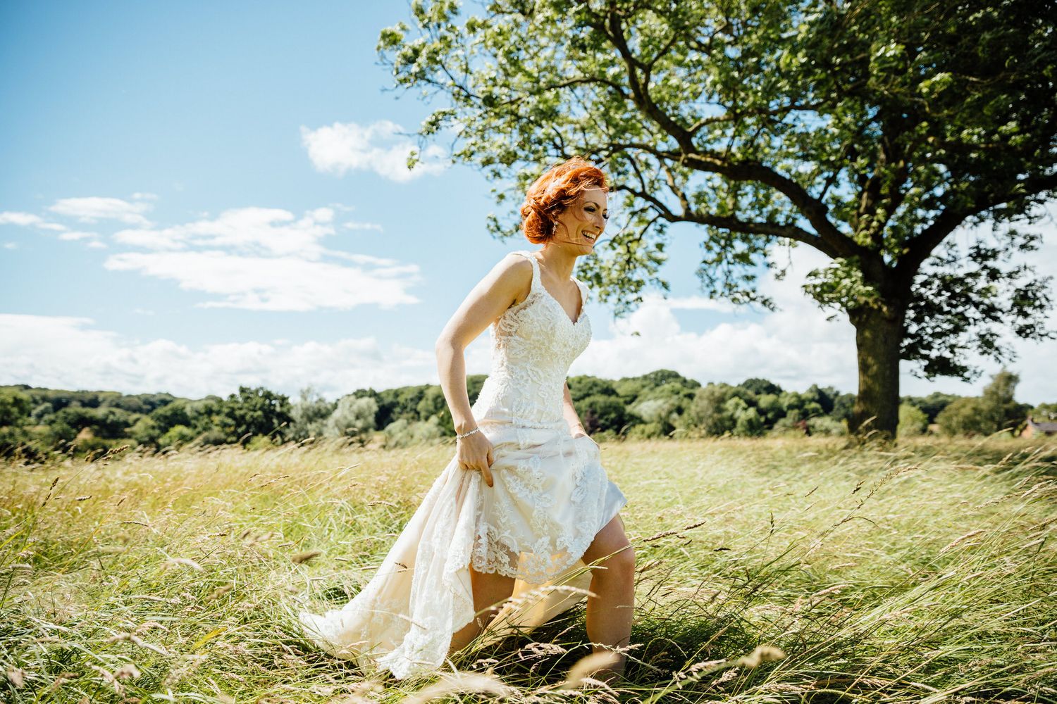 A bride in white dress walking through windswept grass on a sunny day.