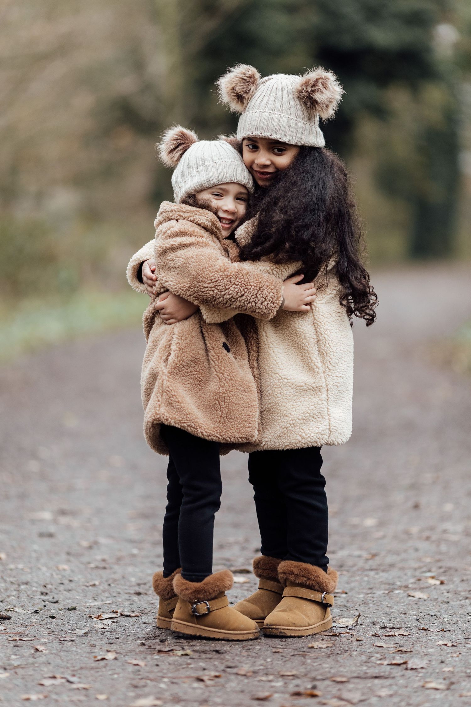 Two people in matching cozy winter outfits with brown boots and knit hats embrace on a forest path.
