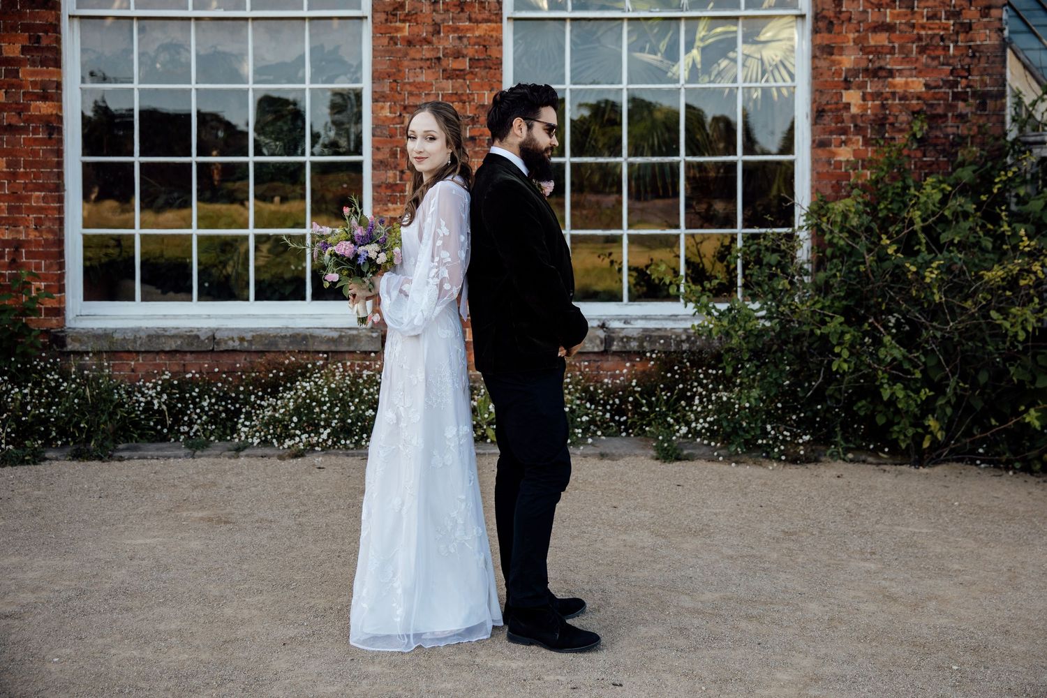 Couple in wedding attire walking along a red brick building with large windows.