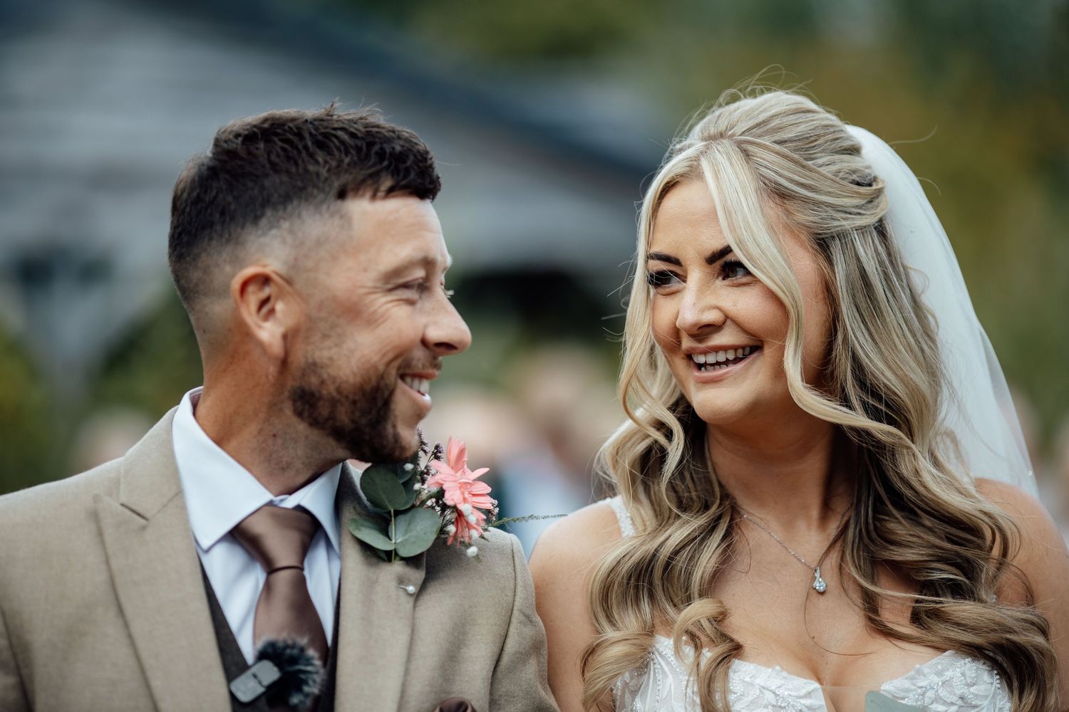 Smiling bride and groom share a joyful moment, bride in lace dress with flowing hair, groom in tan suit with pink boutonniere.