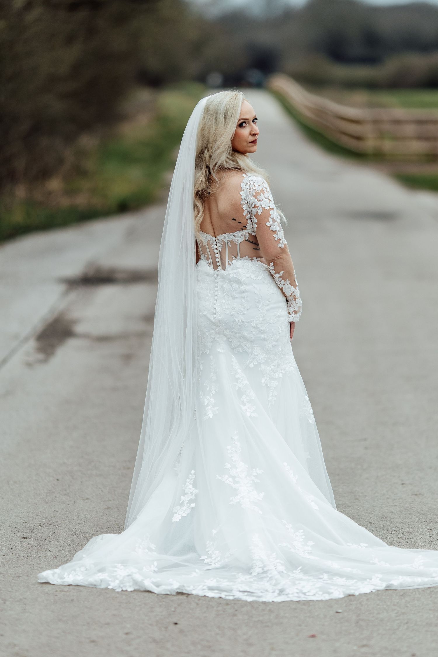 Bride in lace long sleeve wedding gown with cathedral veil poses on a country road.