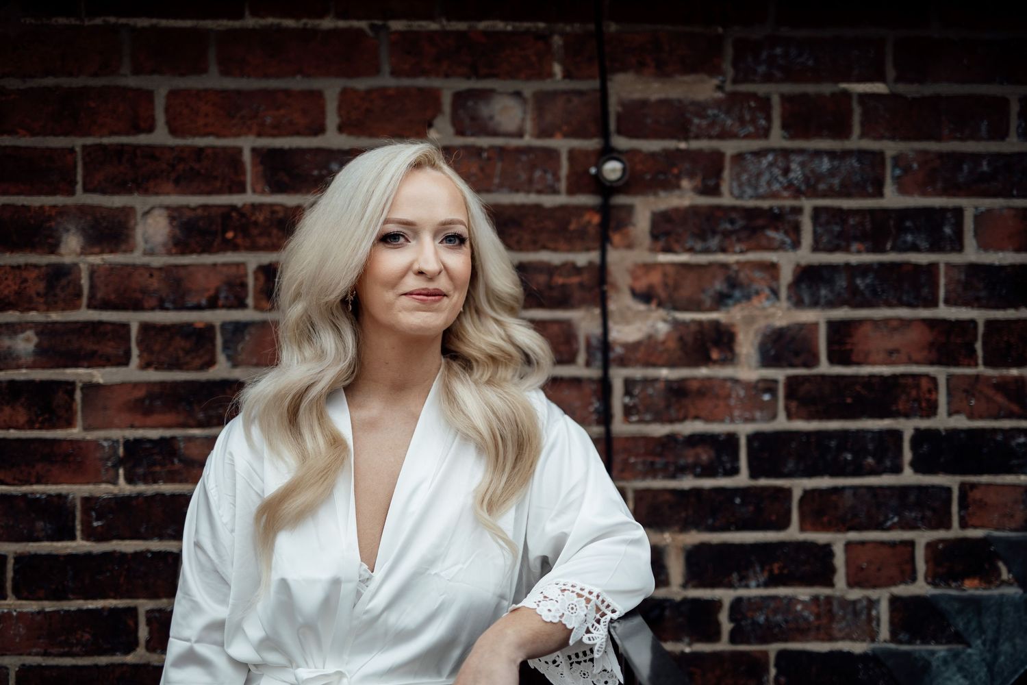 Blonde bride in a white lace robe posing gracefully against a rustic red brick wall.